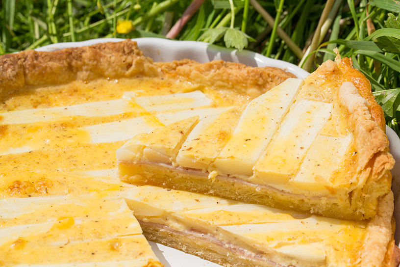 Quiche met asperges en ham aan (taart)stukken op een bord in het gras in de zon